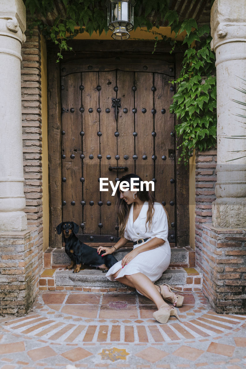 Smiling woman playing with dog while sitting at house doorway