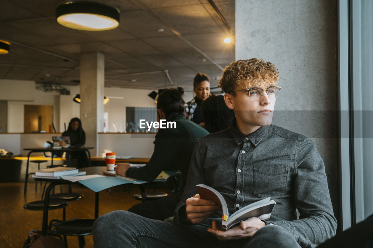 Thoughtful young blond man sitting with book in university cafeteria