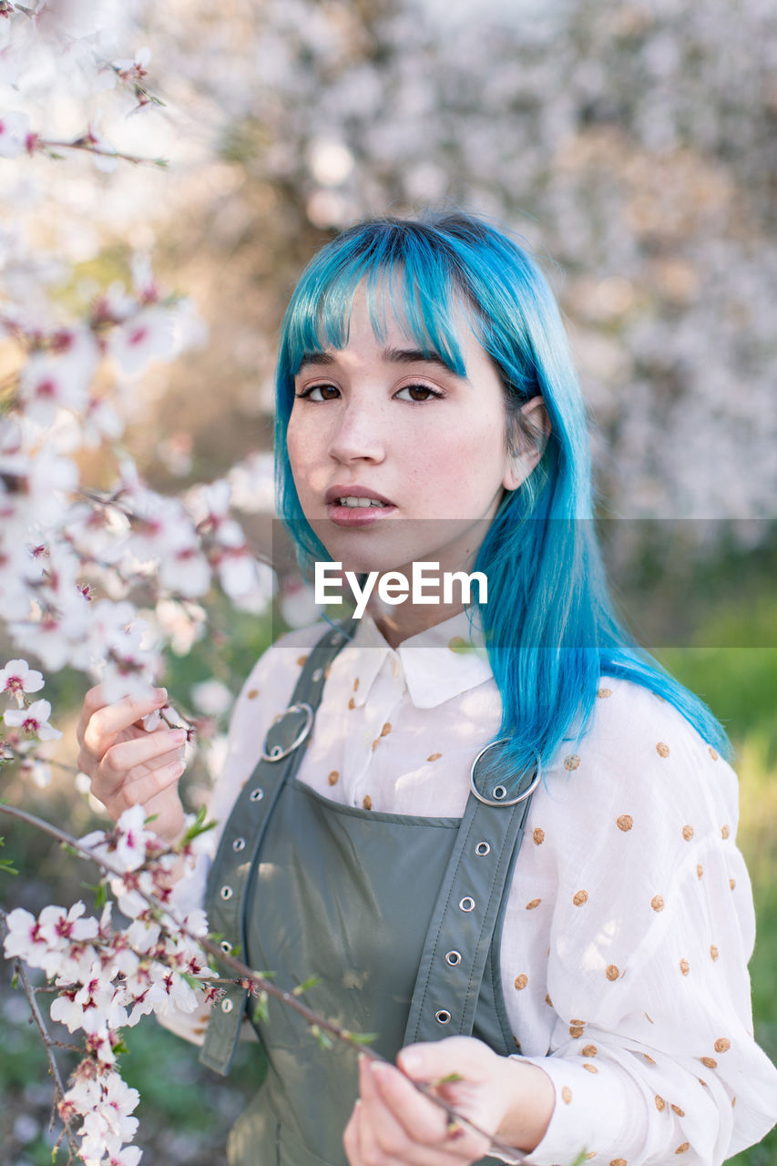 Young stylish female with long blue hair looking at camera wearing trendy overall enjoying blooming tree while standing in spring garden