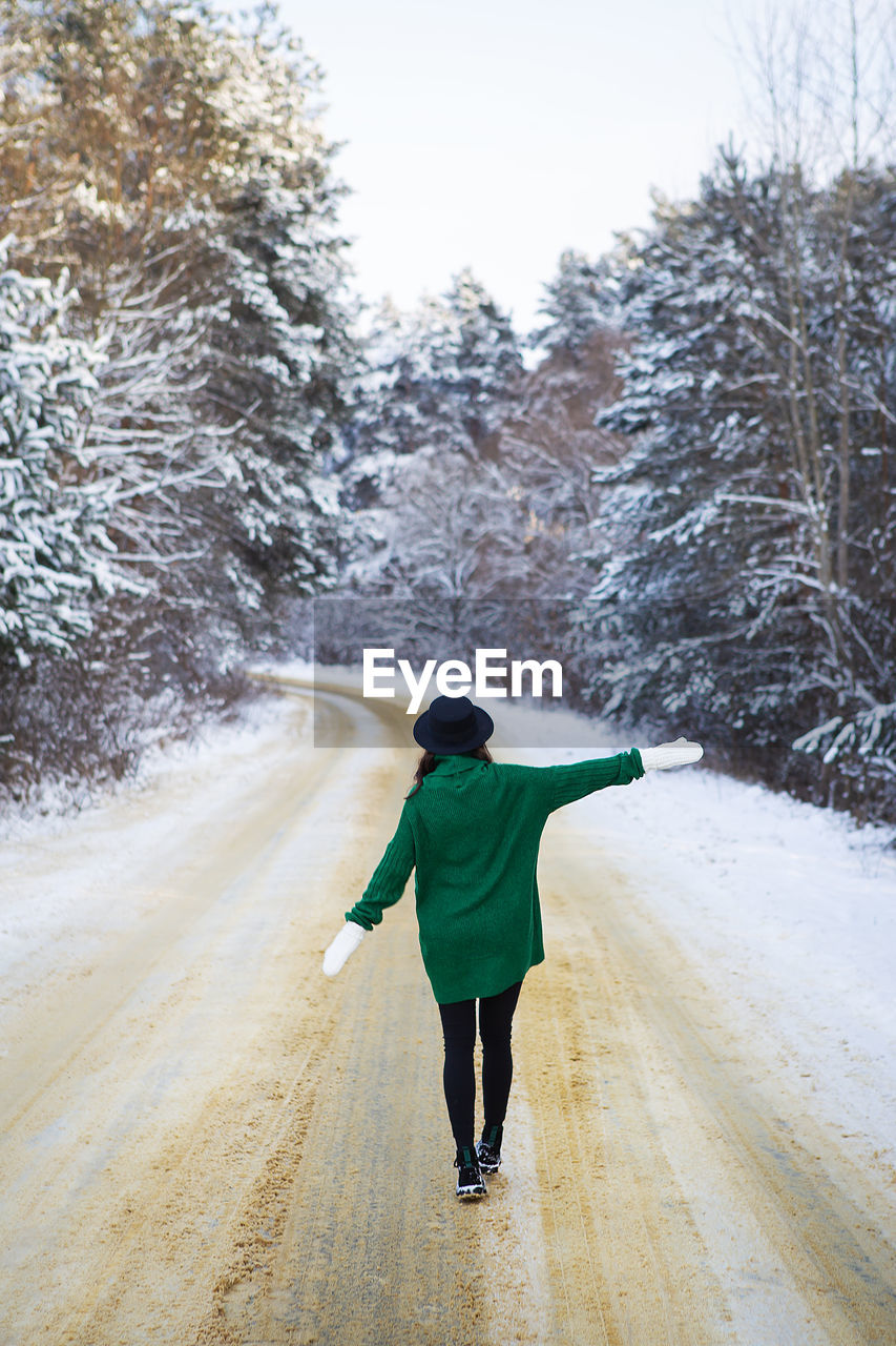 A young girl in a green sweater and hat walks in the middle of a snowy road in a thick pine forest.