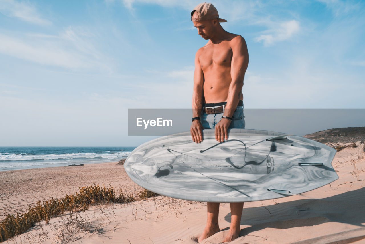 FULL LENGTH OF SHIRTLESS MAN STANDING ON BEACH AGAINST SEA