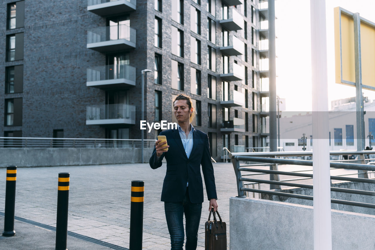 Businessman with briefcase using mobile phone while walking in city