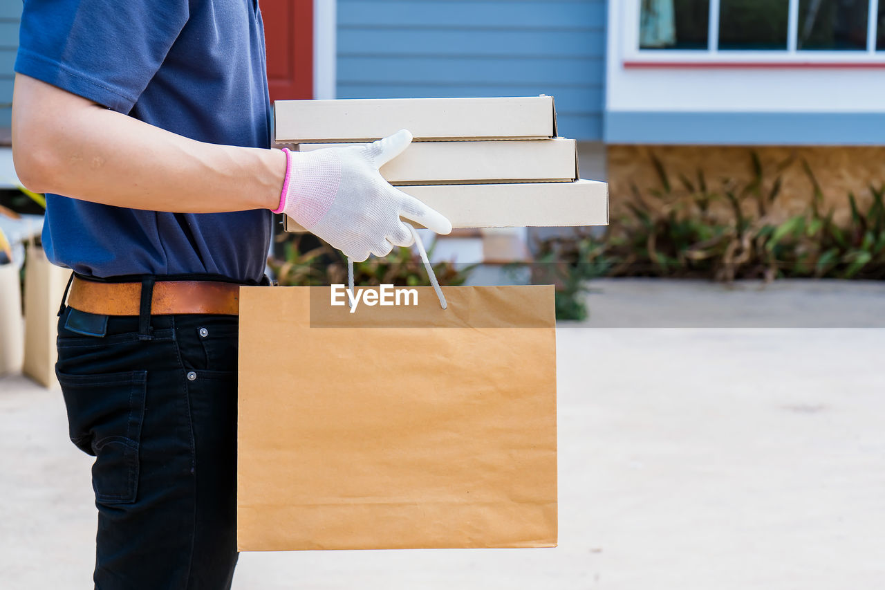 A male delivery man holds a box of goods and food for a customer in front of the house.