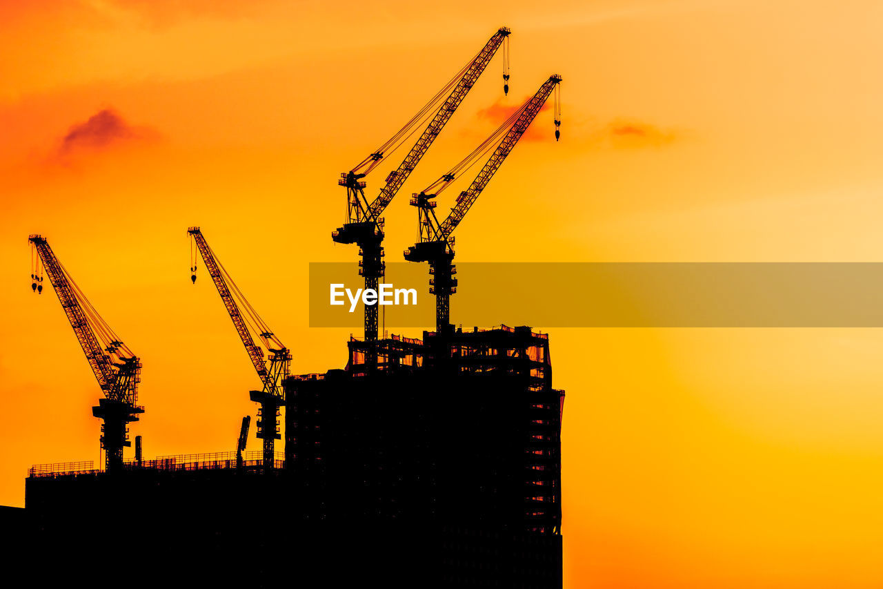 Low angle view of silhouette cranes at construction site against sky during sunset