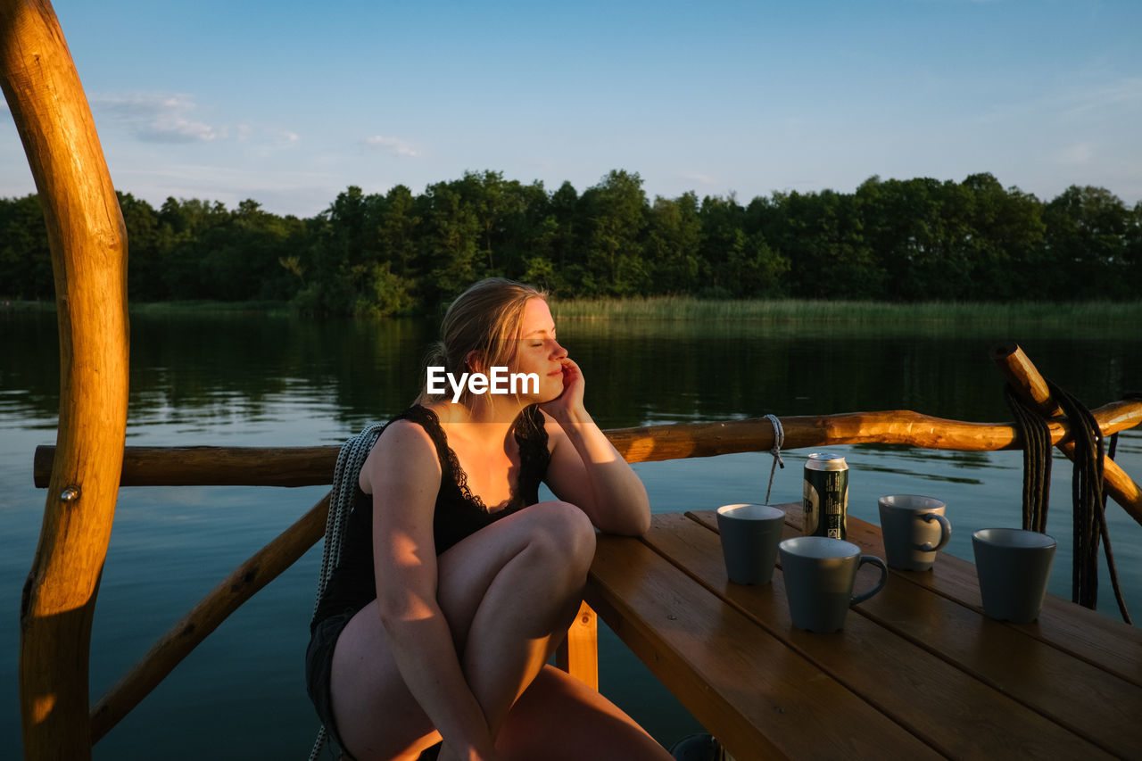 Young woman with eyes closed sitting by lake during sunset