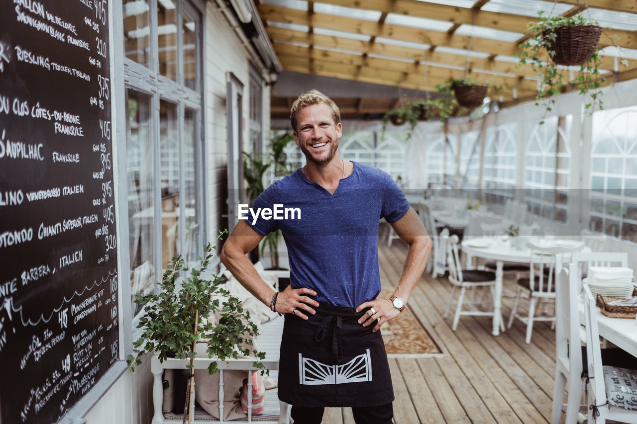 Portrait of smiling male owner with hand on hip standing in restaurant