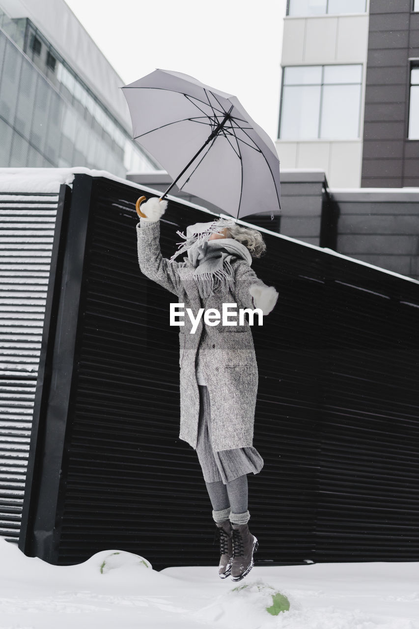 Cheerful woman with umbrella jumping on snowy footpath