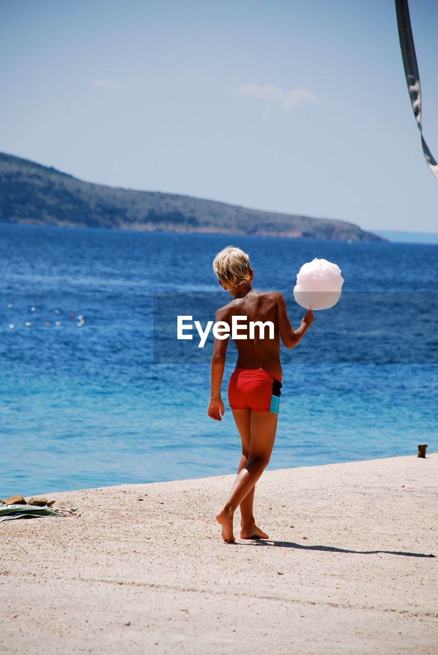 Full length rear view of boy holding cotton candy while walking at beach