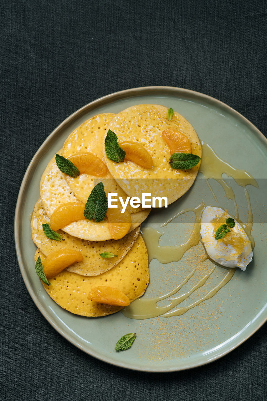 From above of plate with pancakes decorated with slices of tangerine leaves of mint served with sauce and glass of orange juice