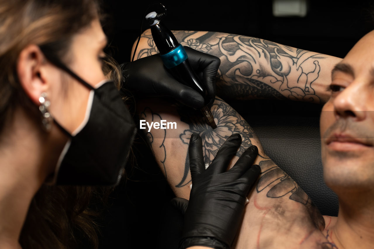 Female tattoo master in mask and with machine making tattoo on arm of male client in dark salon
