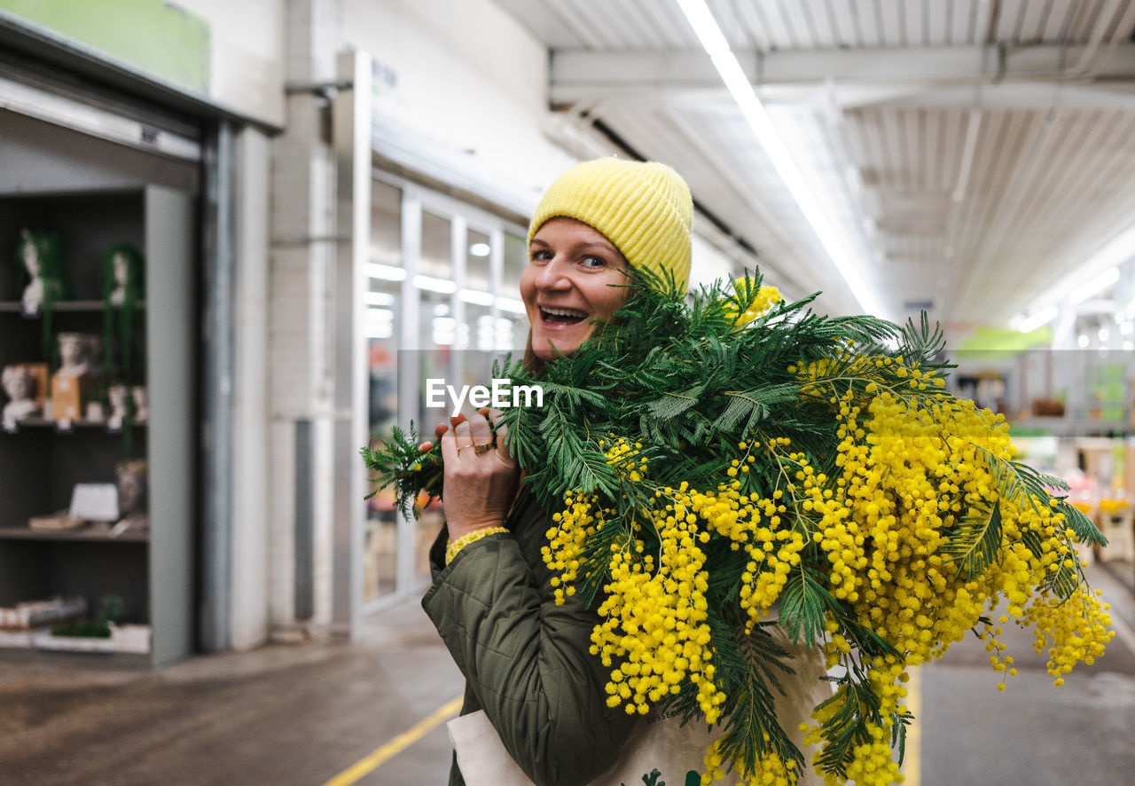 Positive woman in yellow cap and large bouquet of mimosa flowers at flower market