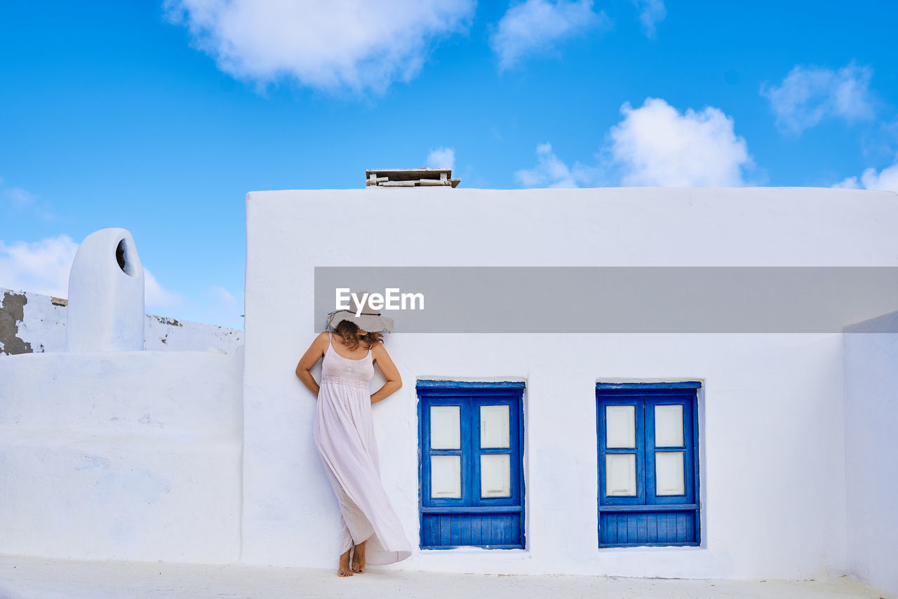 Unrecognizable female in summer clothes covering face with hat while standing outside rural white house with blue window shutters in sunny country