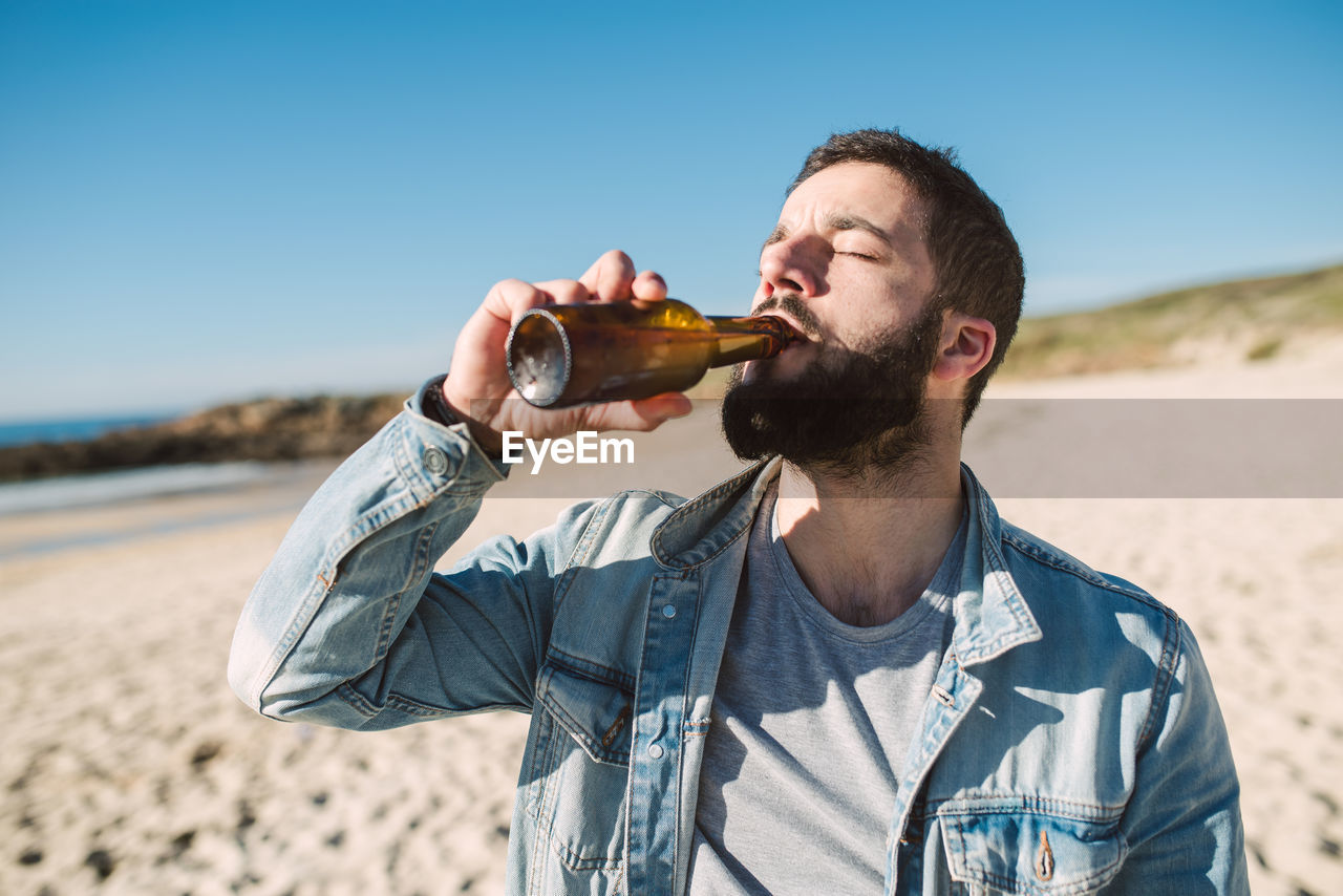 Young man drinking alcoholic drink bottle while standing at beach during sunny day