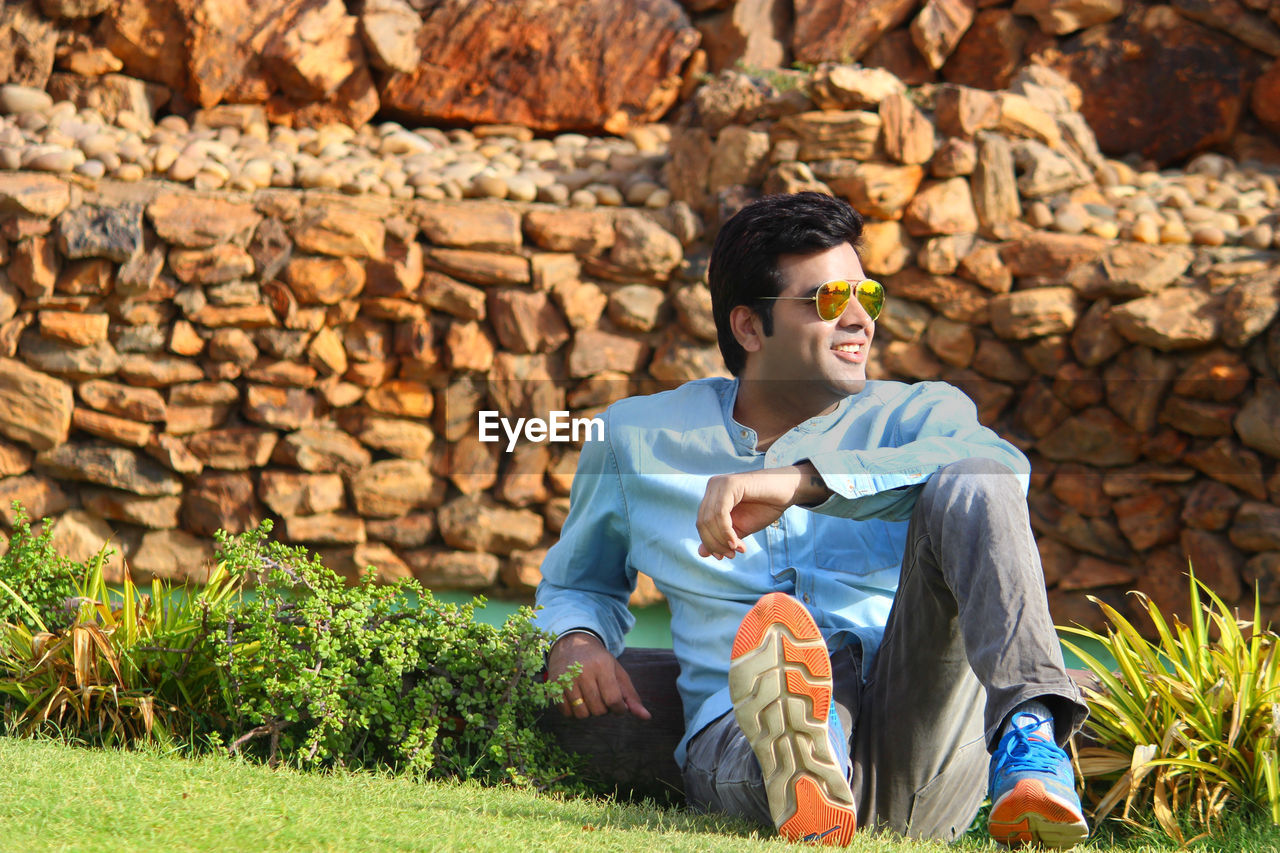 Man wearing sunglasses looking away while sitting against stone wall