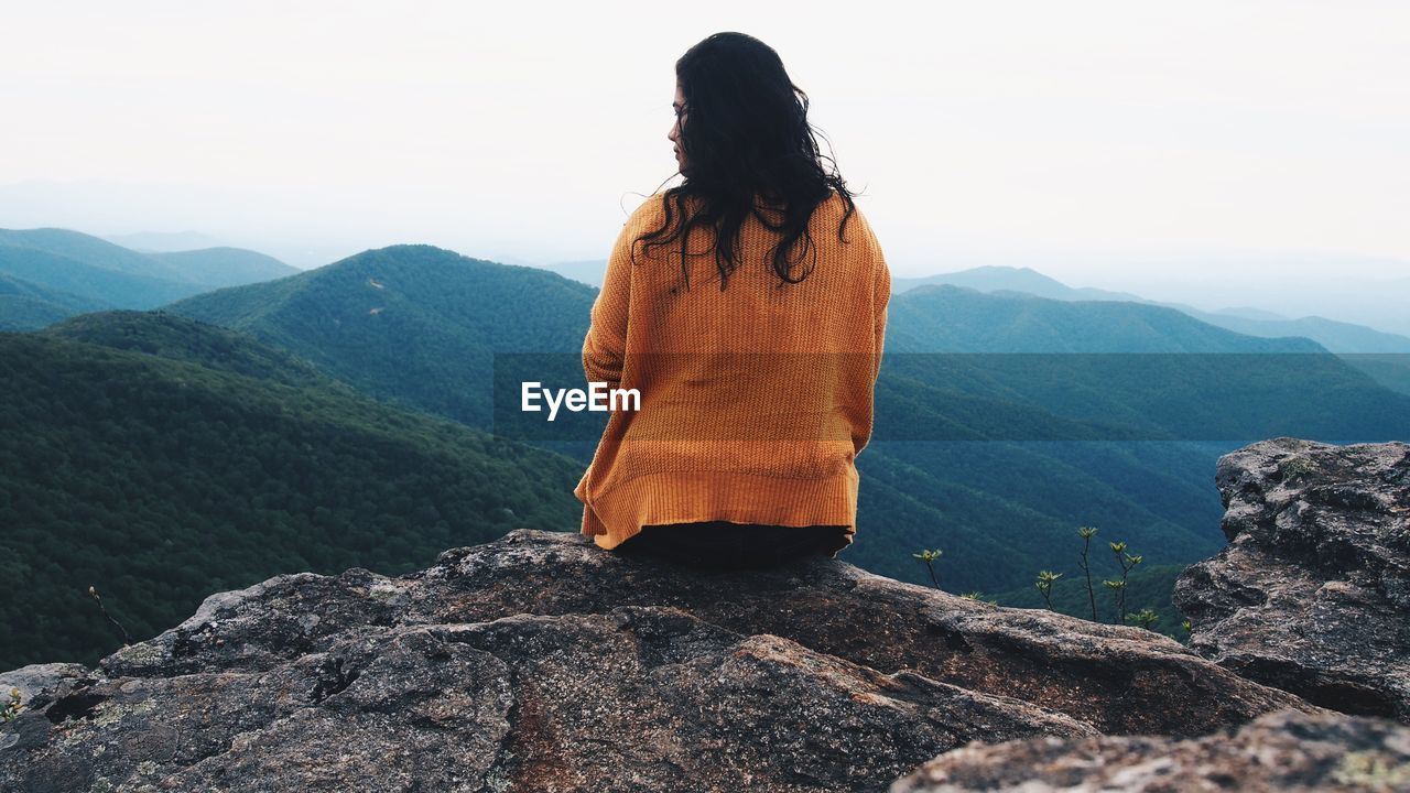 Rear view of woman sitting on cliff against sky