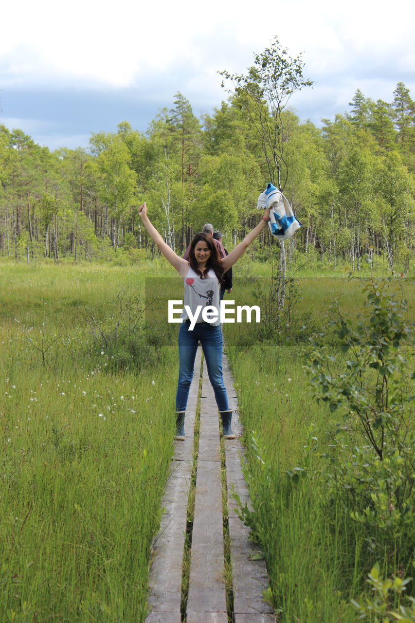 Full length of woman standing with arms raised amidst grassy field against trees