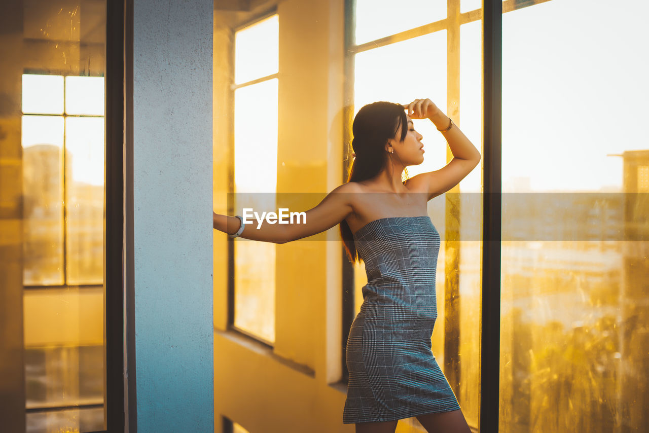 Woman wearing off shoulder dress standing by window at home during sunset