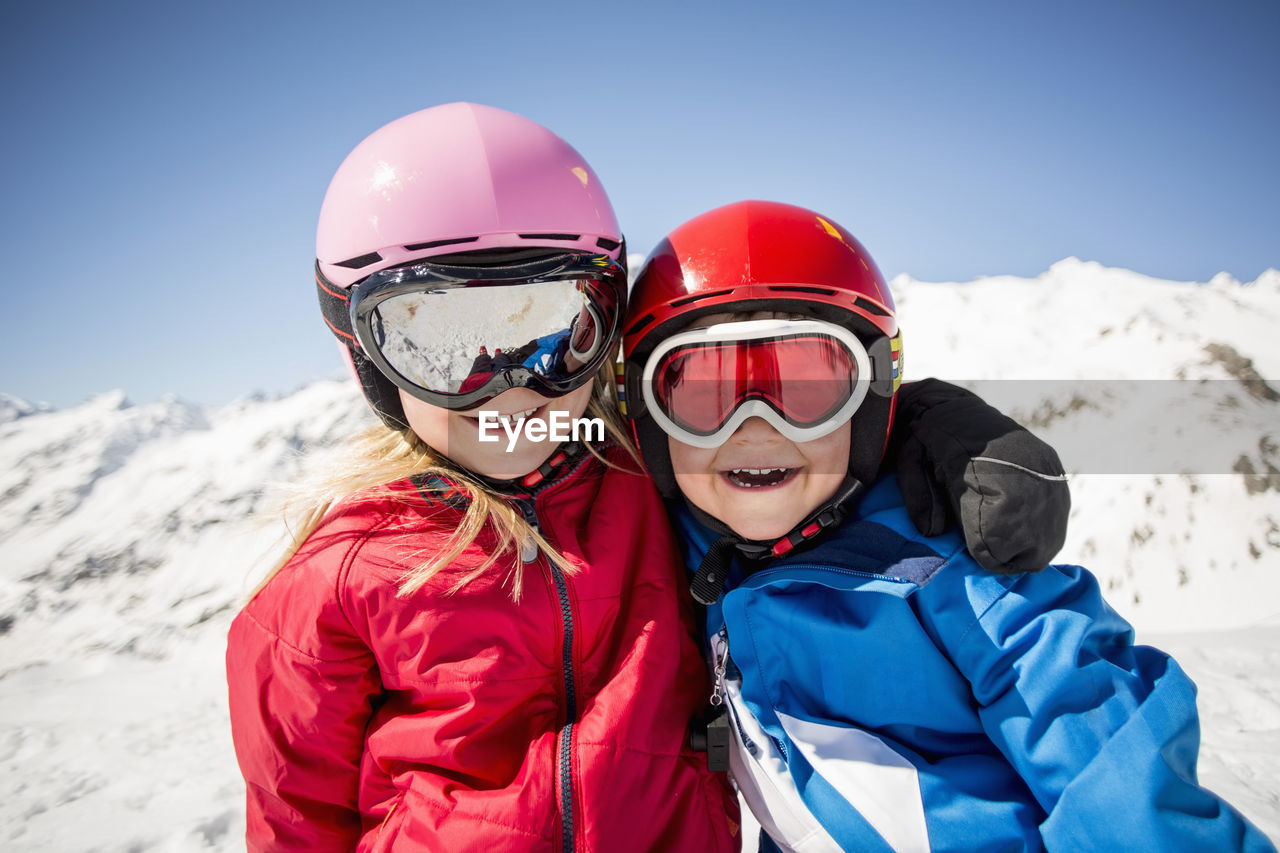 Cheerful siblings in ski-wear standing against snowcapped mountain