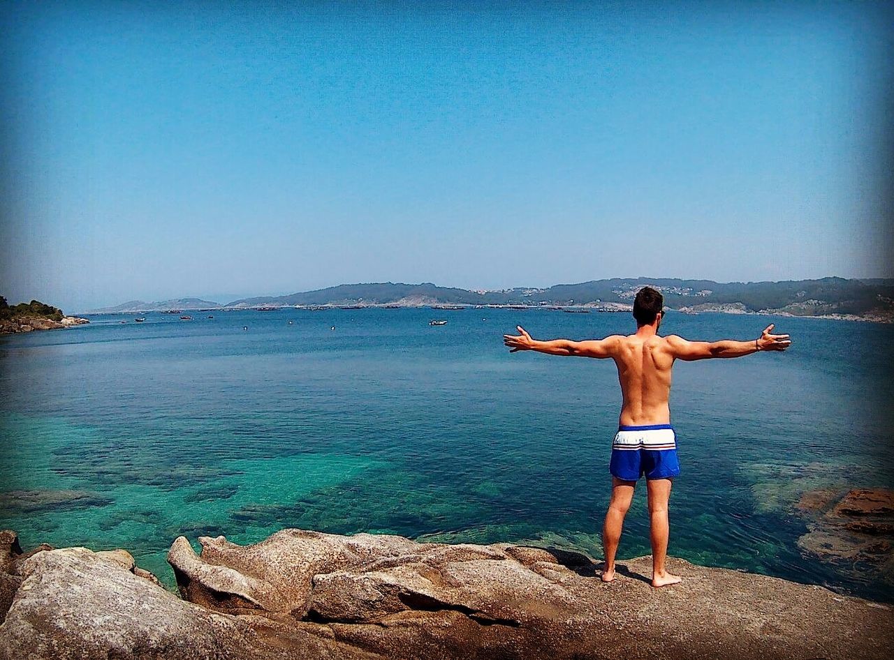 Full length of man with arms outstretched standing on rocks by sea against clear blue sky