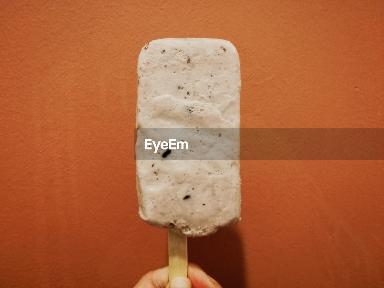 CLOSE-UP OF HAND HOLDING ICE CREAM OVER ORANGE BACKGROUND