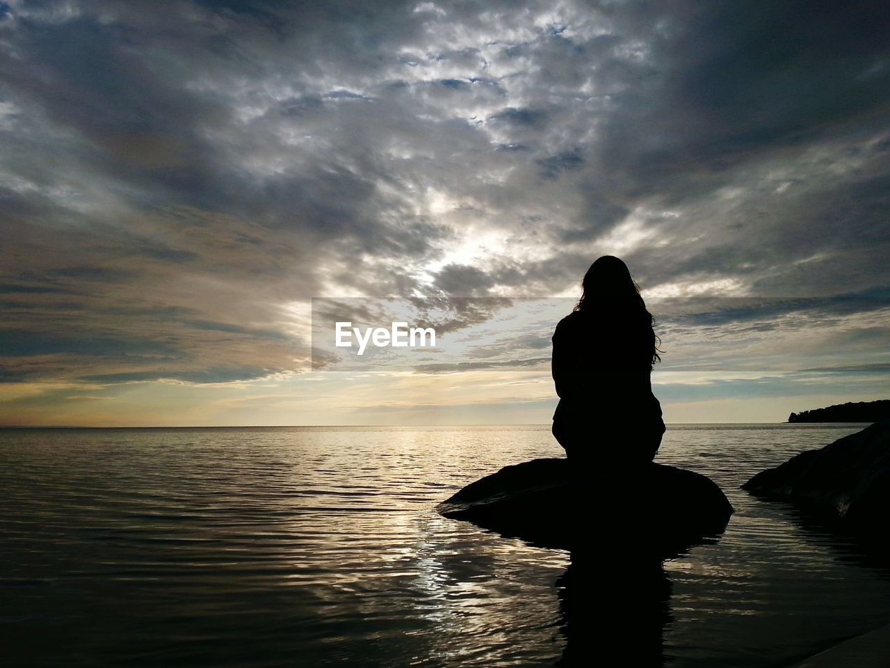 Silhouette woman sitting on top of rock amidst sea against cloudy sky