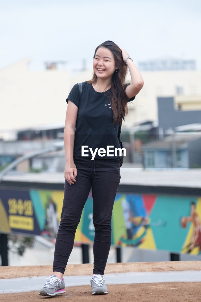 PORTRAIT OF SMILING YOUNG WOMAN STANDING AGAINST BLURRED BACKGROUND