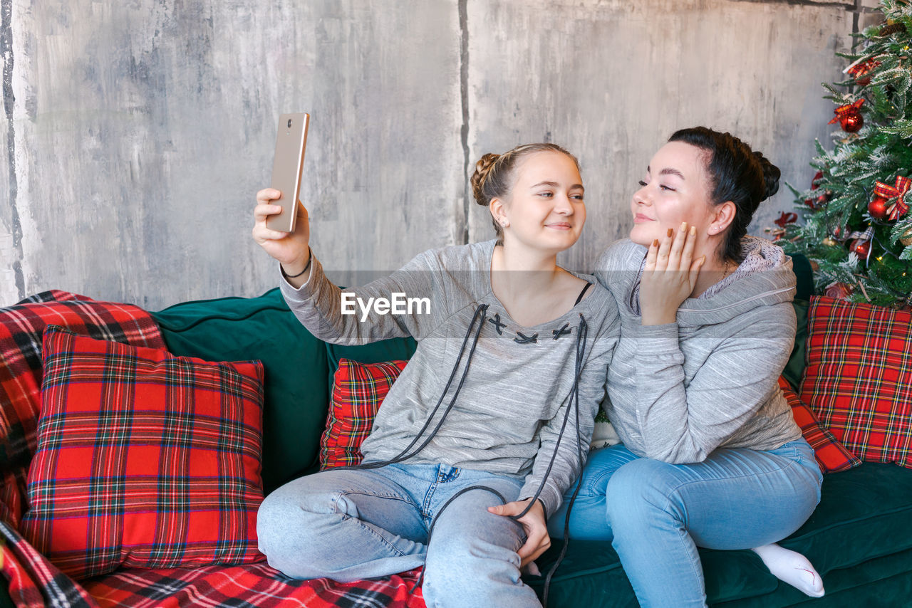 Two pretty young sisters in gray sweatshirts and blue jeans sit on couch near