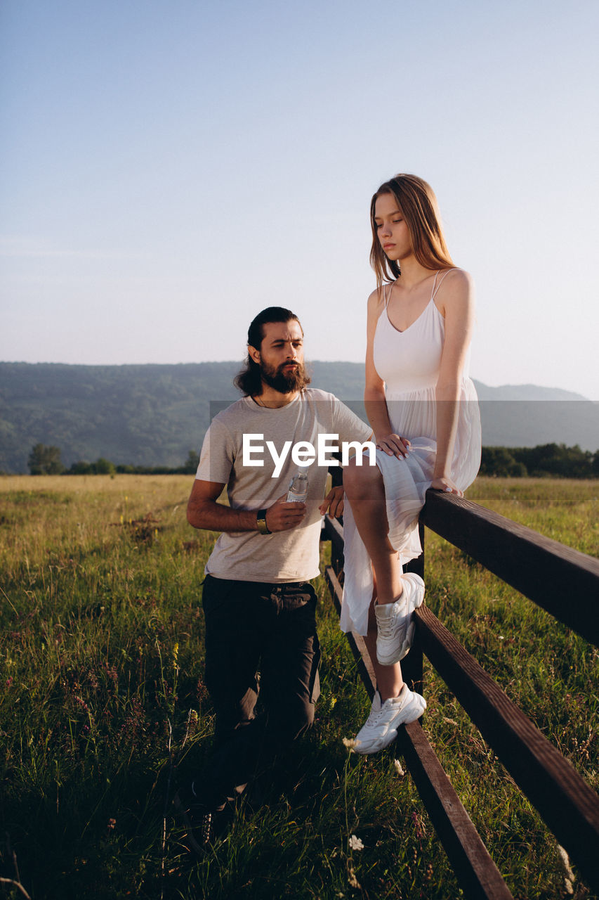 Bearded man standing by woman sitting on railing against clear sky