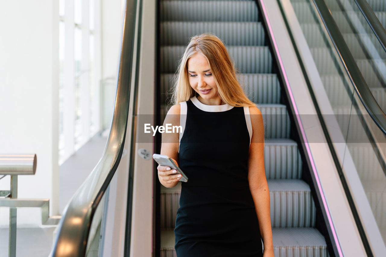 Blond female in black dress using smartphone and looking at screen with smile while standing near moving stairs in modern mall