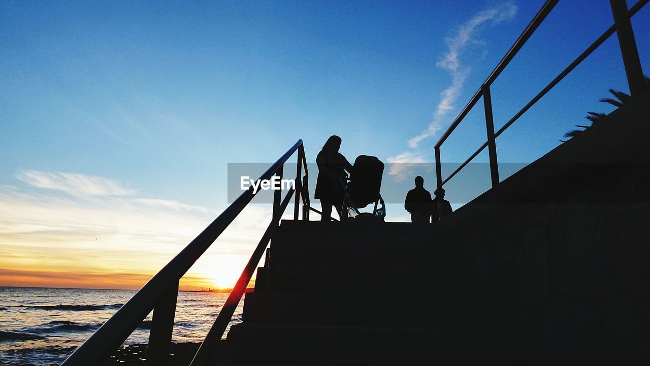 Low angle view of silhouette people standing on railing by sea