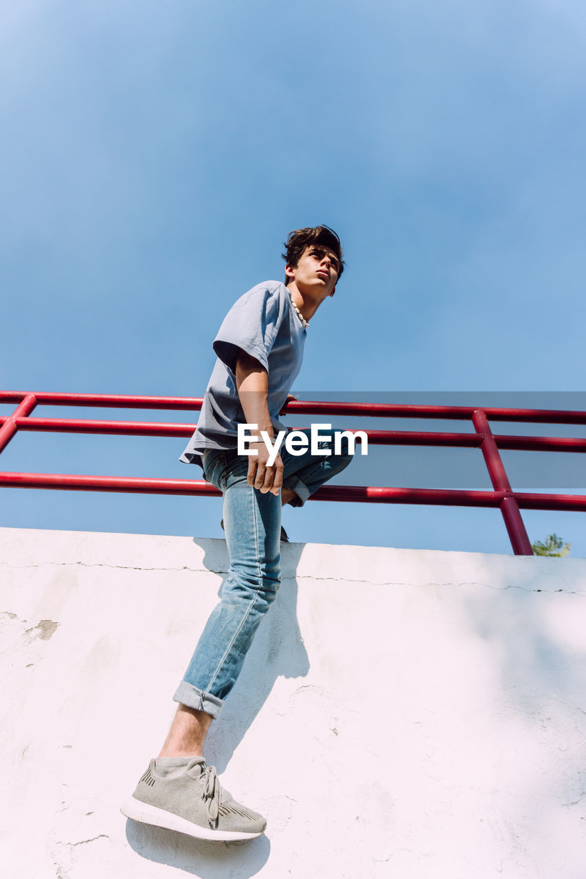 From below side view of of brave male hanging looking away over metal fence in street and showing parkour trick against blue sky