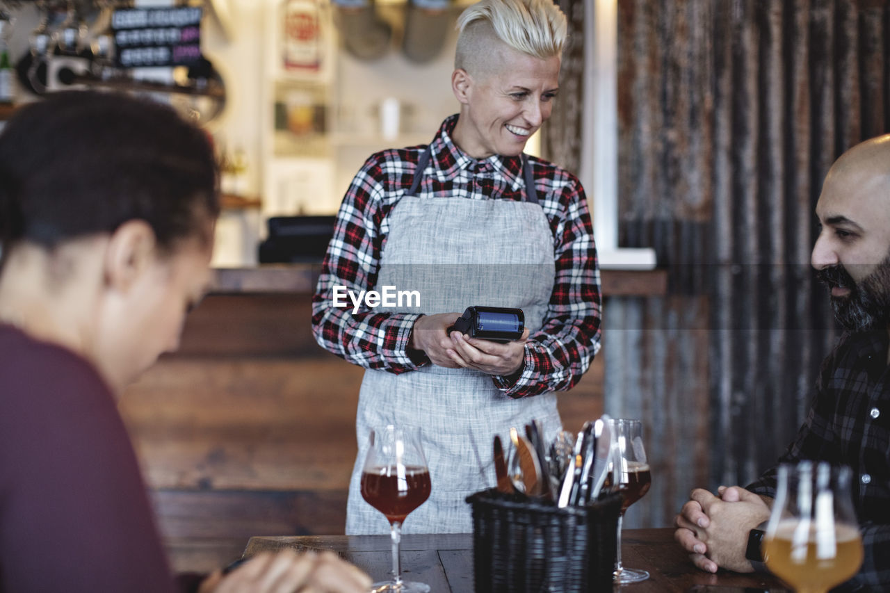 Smiling female bartender holding credit card reader while talking to customers at table in bar