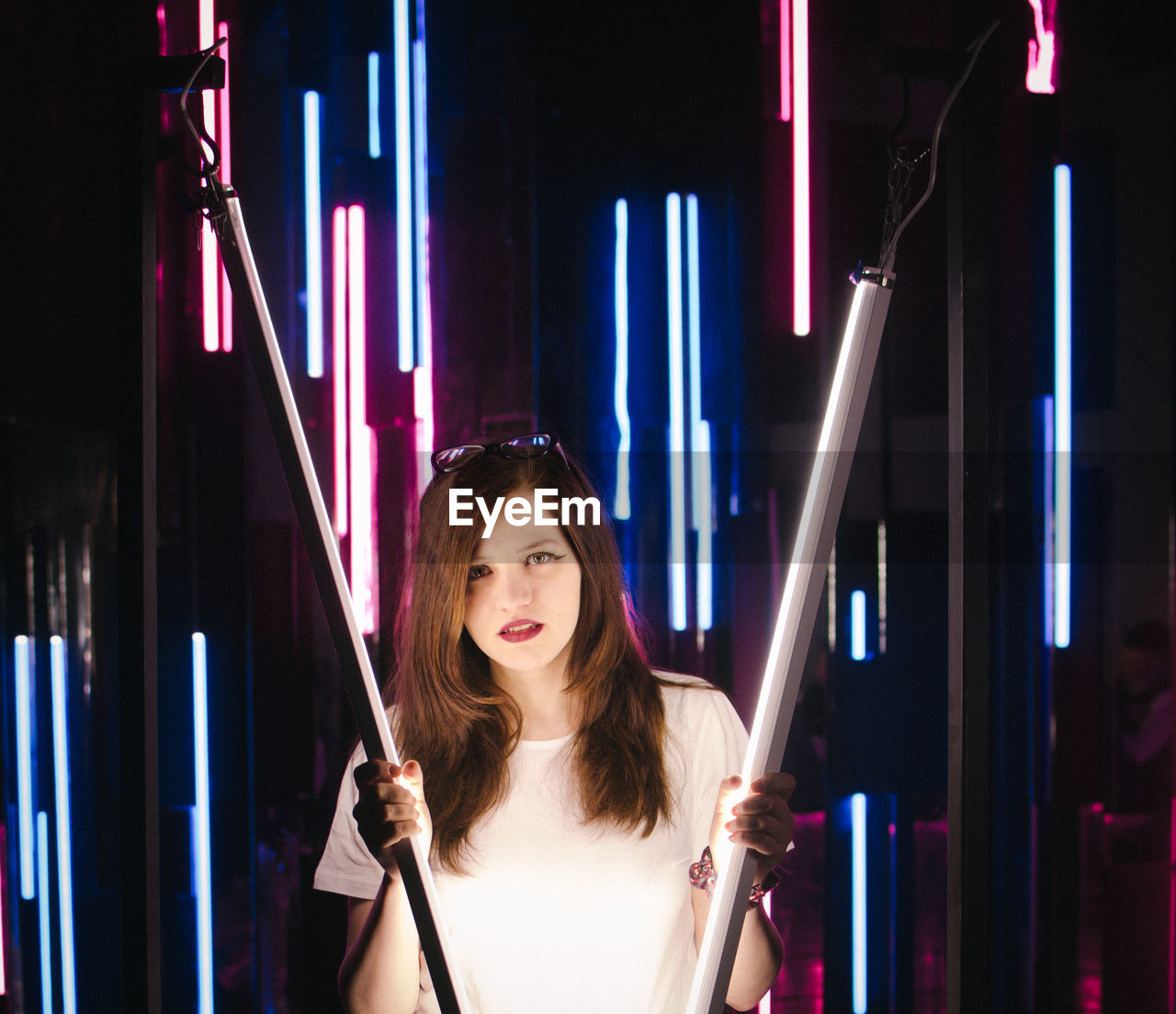 PORTRAIT OF YOUNG WOMAN STANDING AGAINST ILLUMINATED WALL