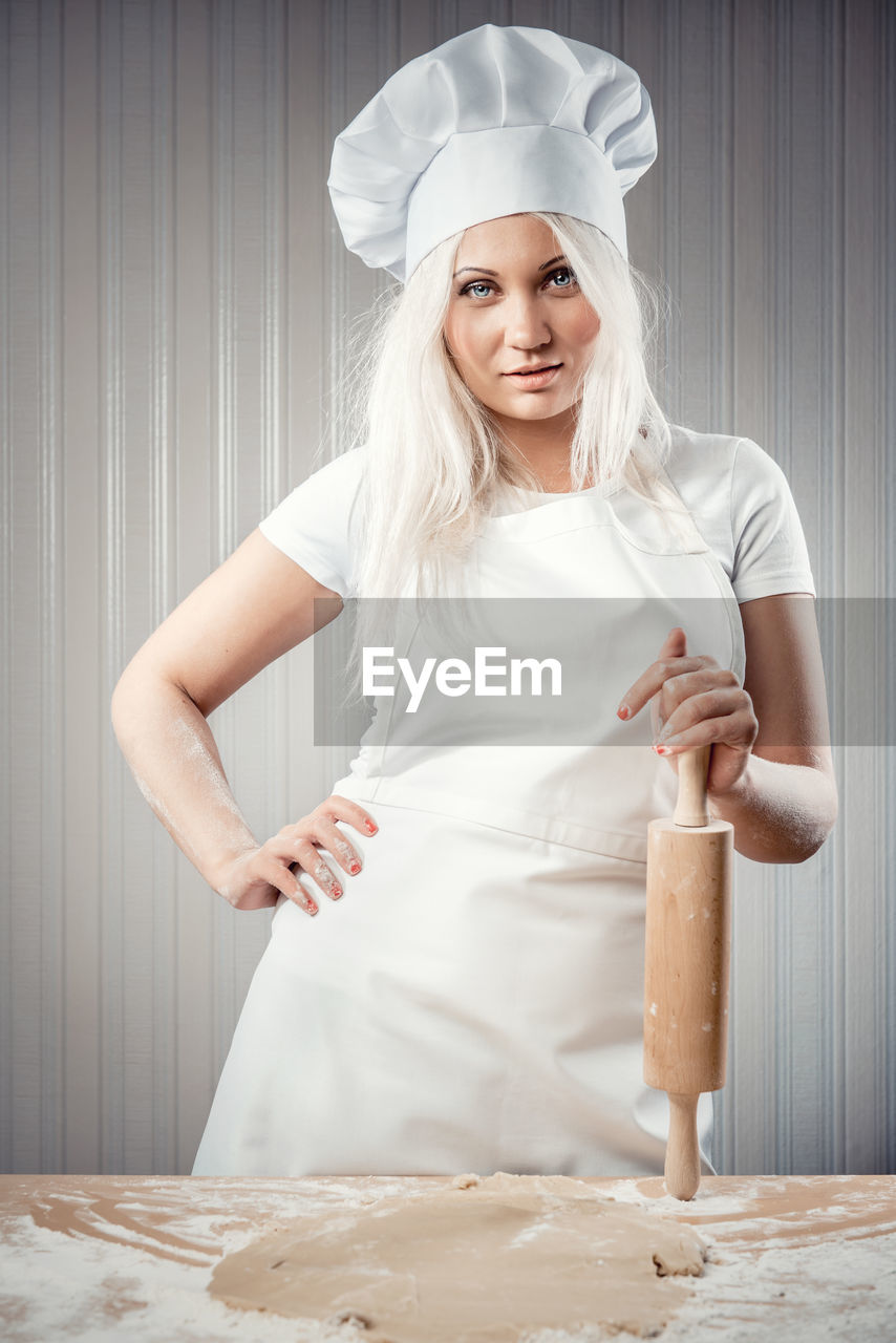 Portrait of young woman holding rolling pin while standing in kitchen
