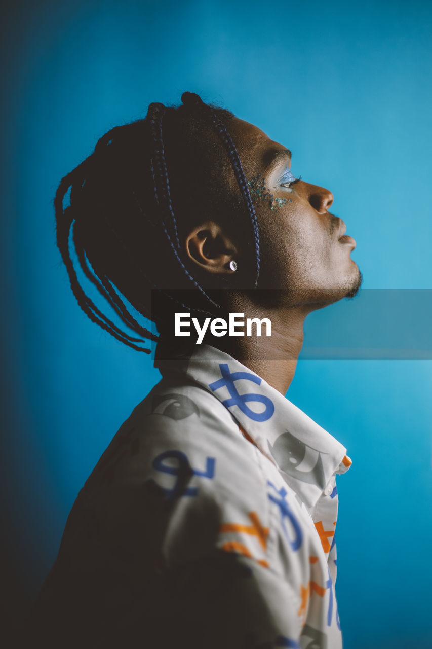 Serious african american male with braided hairstyle and bright makeup looking away on blue background in studio