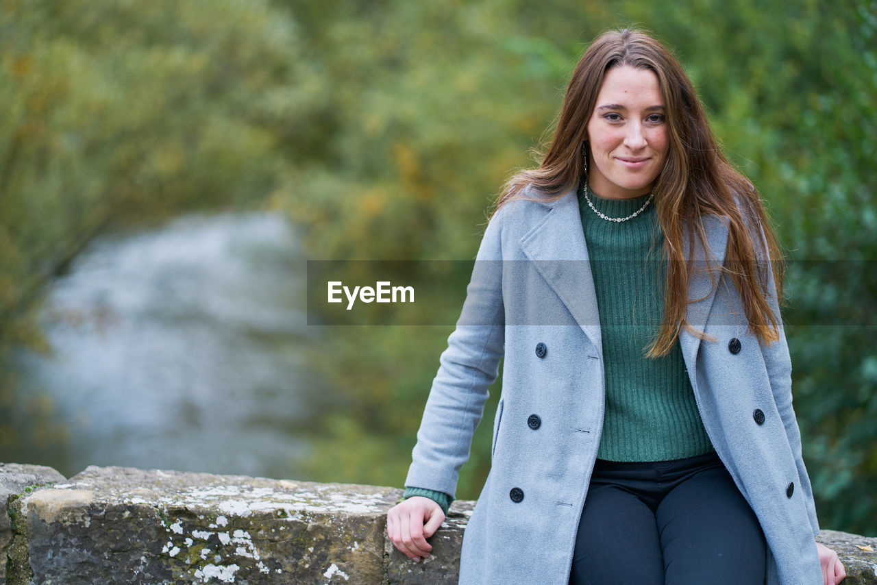 Portrait of a young caucasian woman with brown hair on a stone bridge with the green of the trees in the background. dressed in a gray jacket, green sweater, dark pants and military boots
