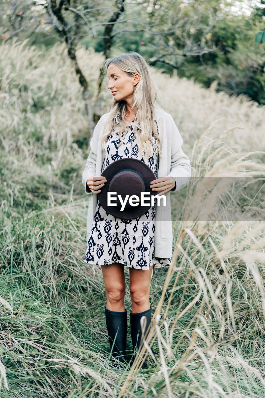 Beautiful pregnant woman stands in the park in autumn and covers her belly with a hat