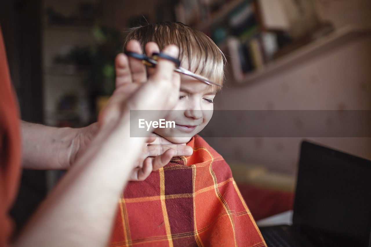 Middle aged father cutting hair to his son by himself at home looking video broadcast on laptop