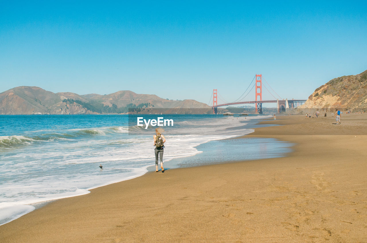 Full length rear view of woman running on beach by golden gate bridge against clear blue sky
