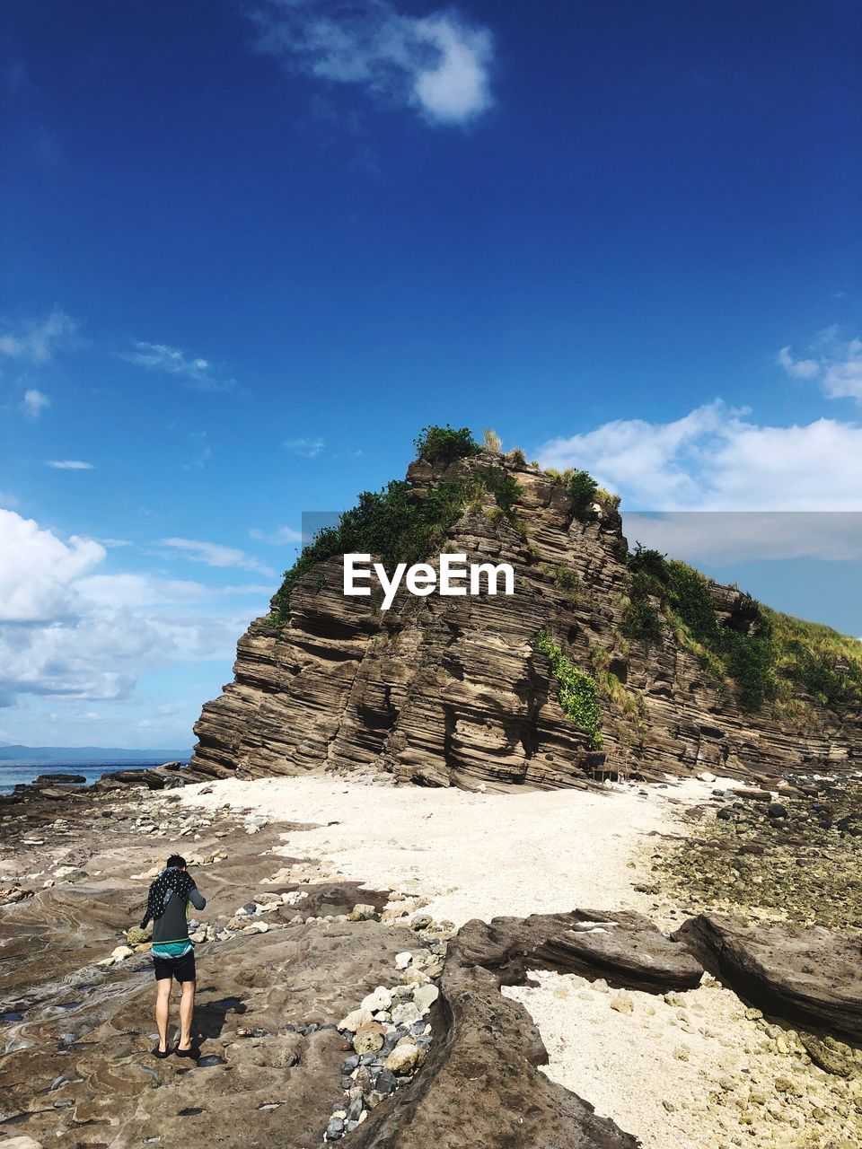 REAR VIEW OF WOMAN STANDING ON ROCK BY MOUNTAIN AGAINST SKY