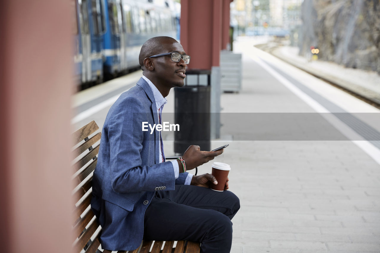 Thoughtful businessman sitting with smart phone and coffee at railroad station