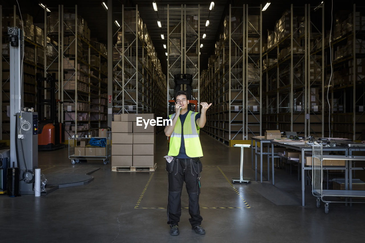 Full length of young male warehouse worker gesturing while using walkie-talkie at storage room