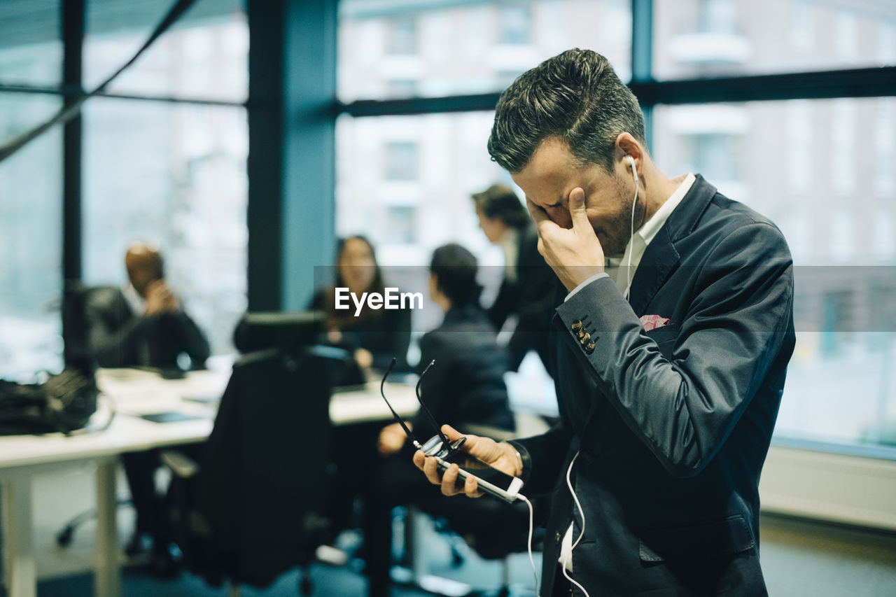 Stressed businessman using phone through headphones while rubbing eyes in office