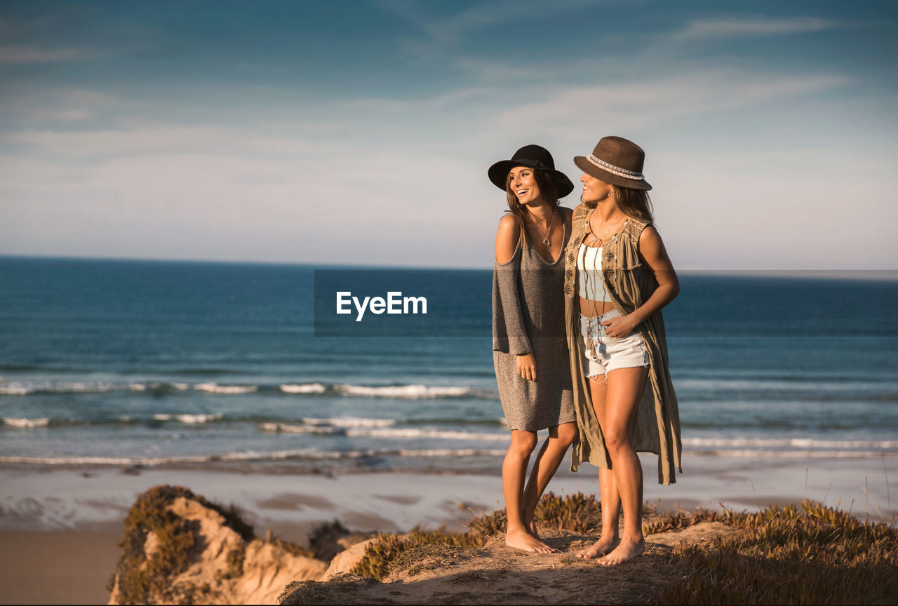 Female friends wearing hats while standing at beach against sky