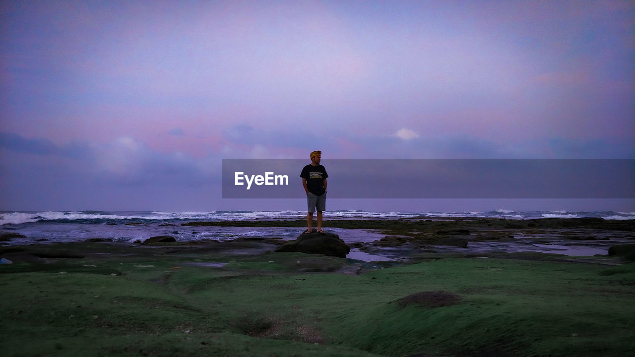 REAR VIEW OF MAN STANDING AT BEACH DURING SUNSET