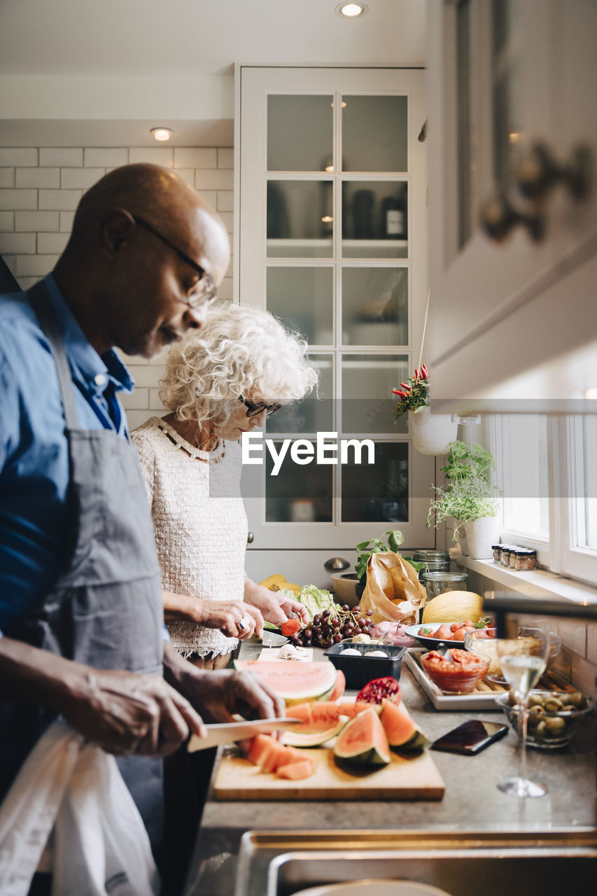 Multi-ethnic senior friends cutting fruits at counter while preparing dinner in kitchen