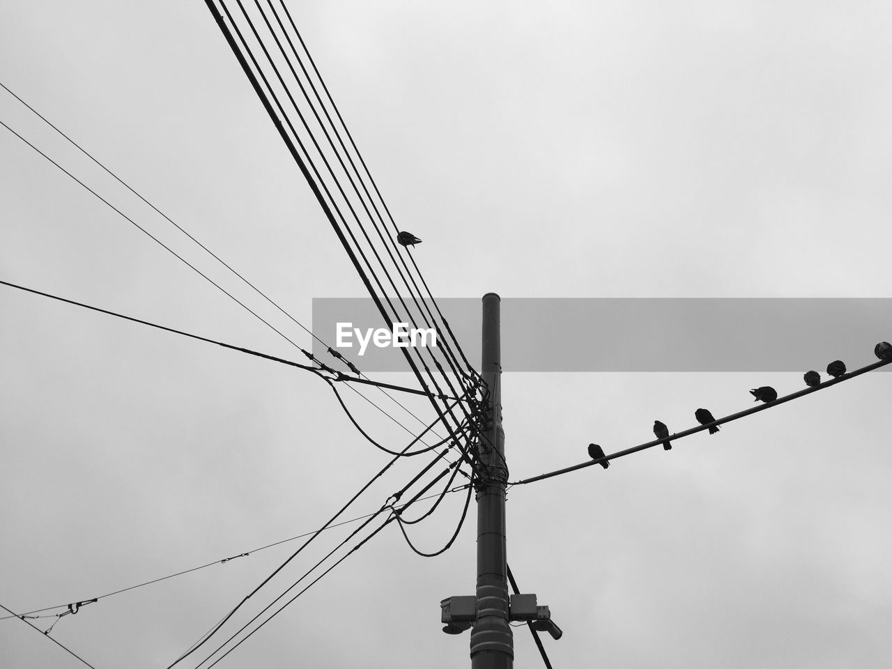 Low angle view of electricity pole and cables against cloudy sky