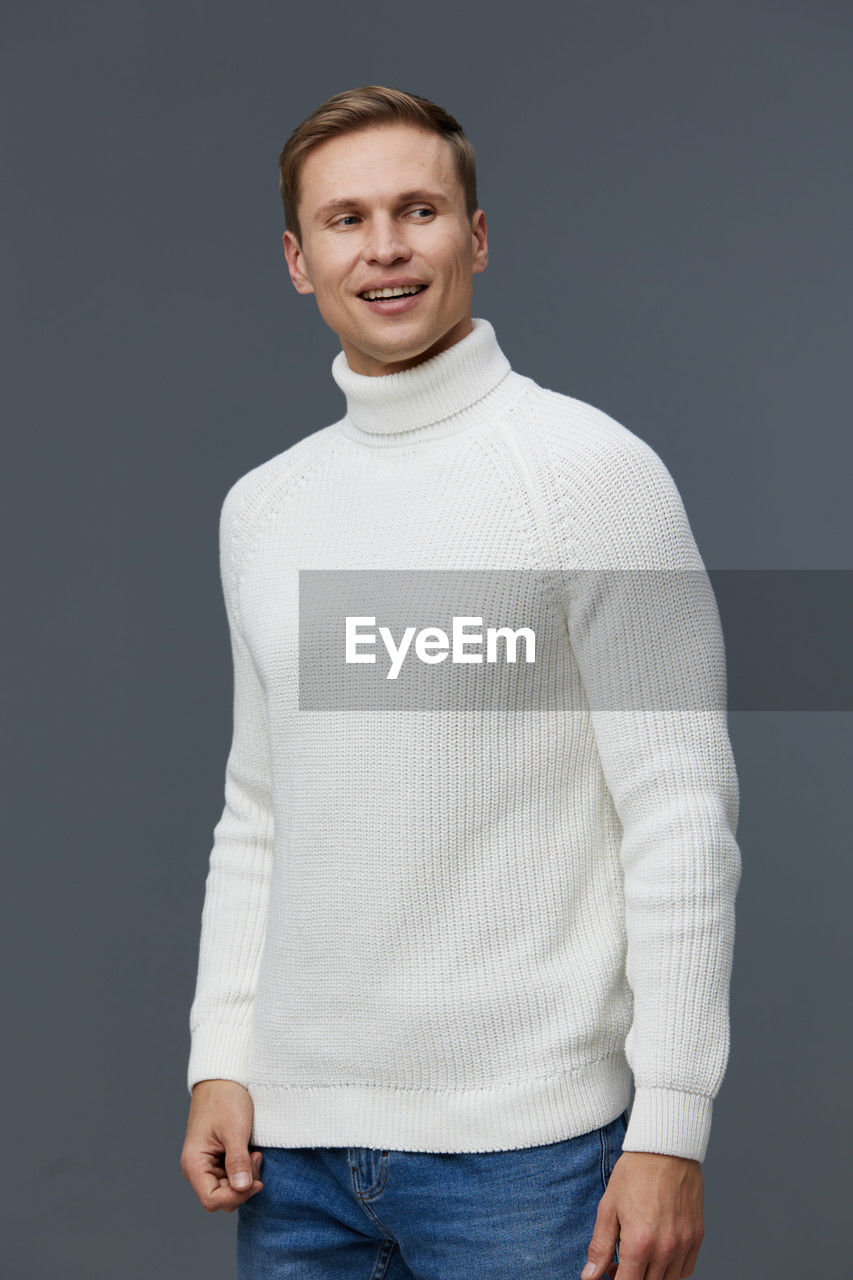 portrait, studio shot, looking at camera, sleeve, one person, smiling, indoors, clothing, casual clothing, gray, sweater, happiness, outerwear, t-shirt, standing, adult, emotion, young adult, pocket, gray background, long-sleeved t-shirt, jeans, hands in pockets, three quarter length, men, white, front view, collar, cheerful, waist up, fashion, brown hair, photo shoot, teeth, smile, cut out, relaxation, wool, hairstyle