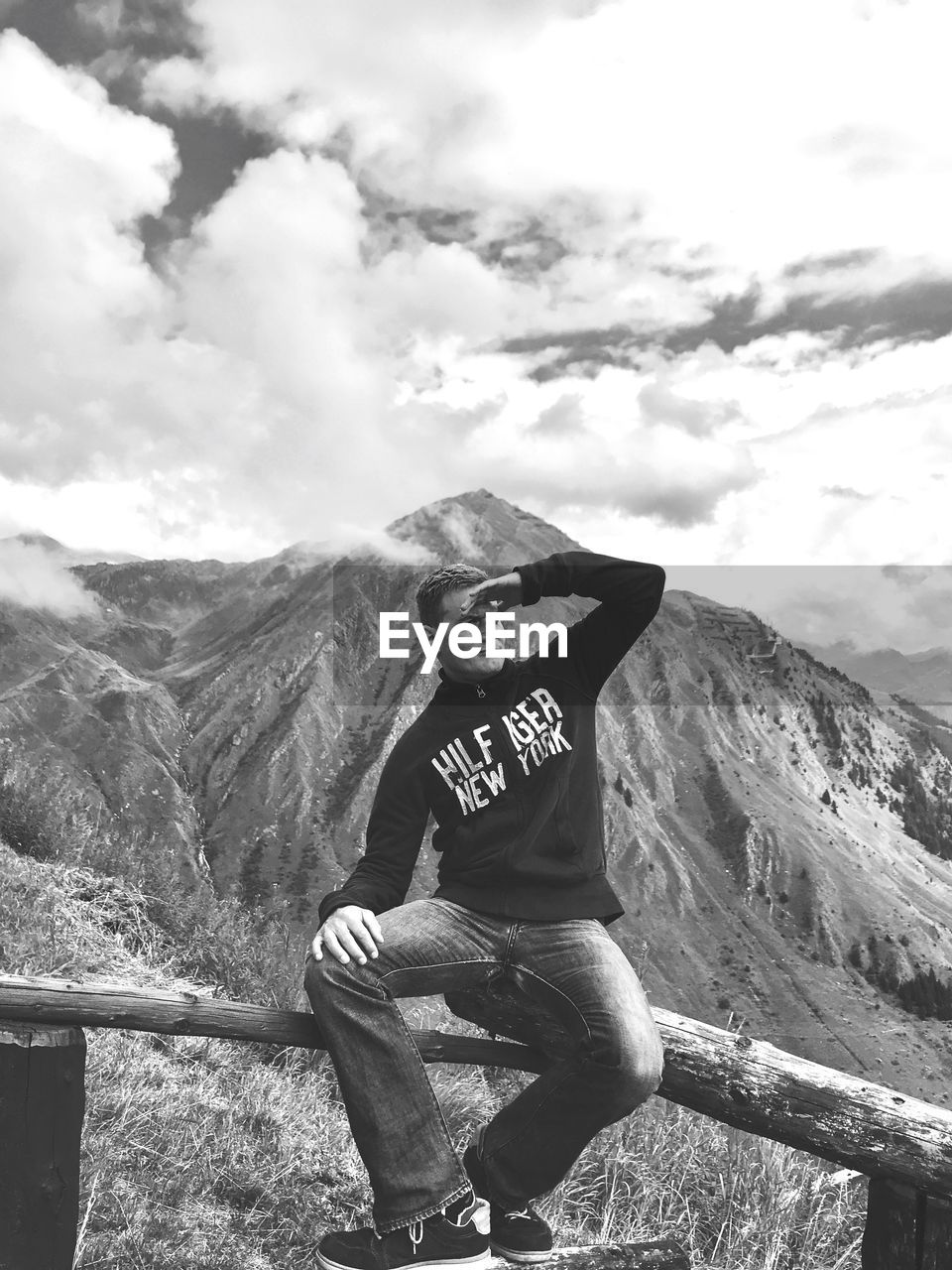 Man sitting on railing with mountains in background against cloudy sky