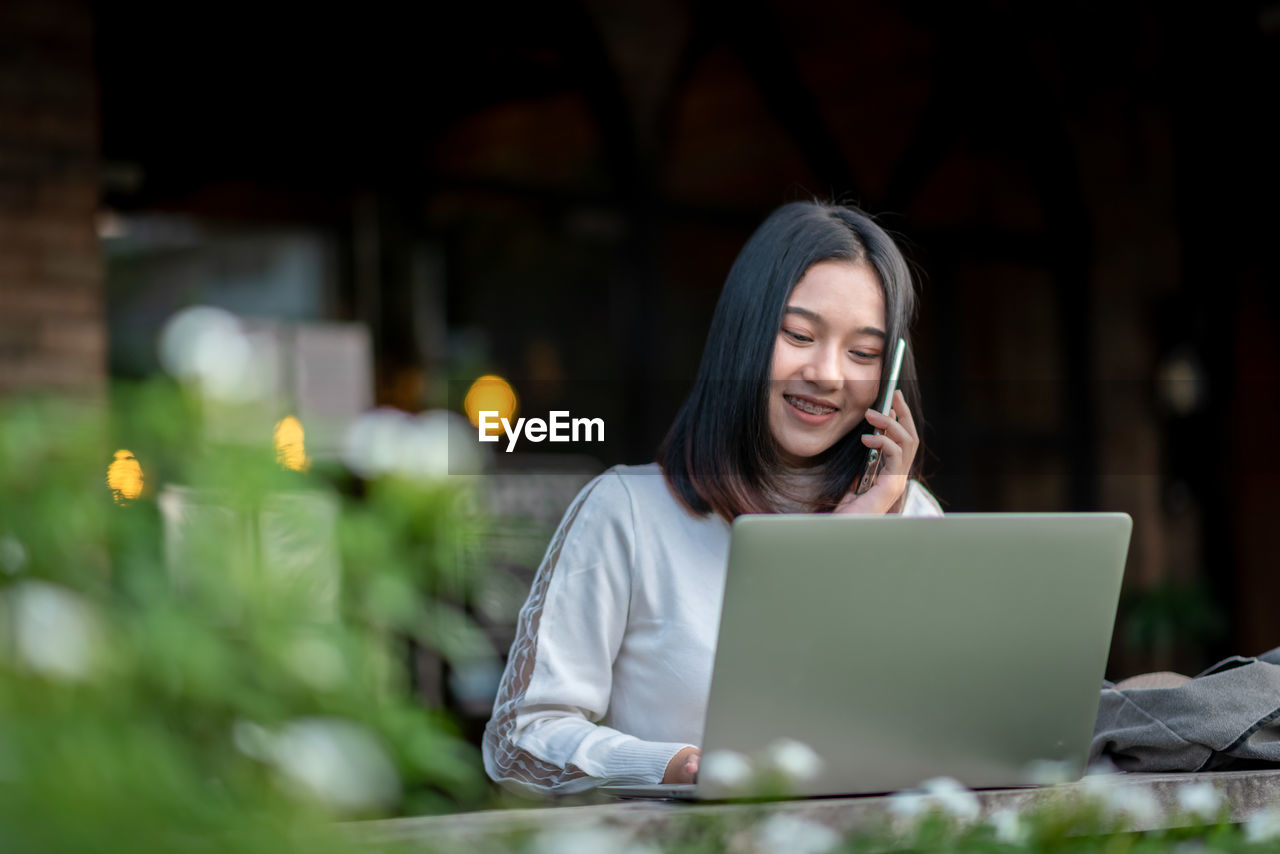 PORTRAIT OF A SMILING YOUNG WOMAN USING MOBILE PHONE IN PARK